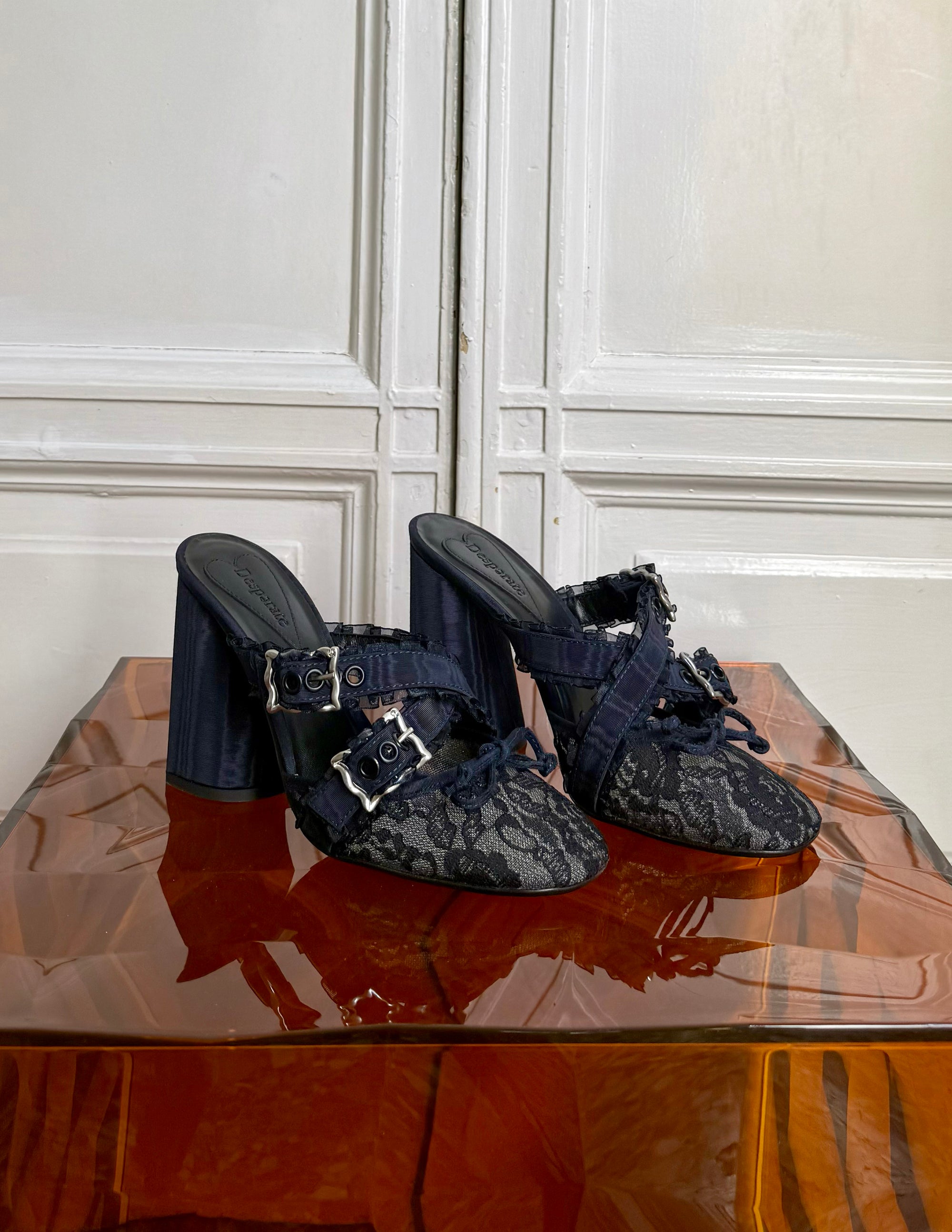Pair of black lace high-heeled shoes on a reflective surface with a white paneled wall background.
