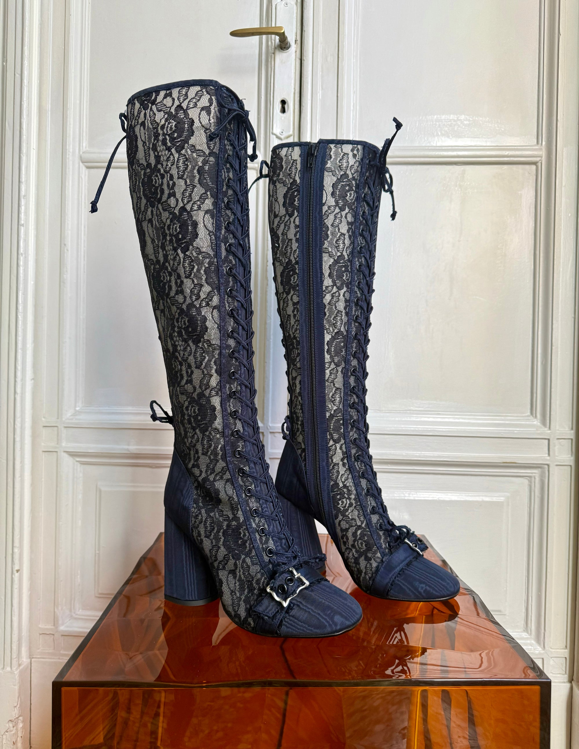 Pair of navy blue lace-up knee-high boots on a wooden platform with a white paneled wall background.