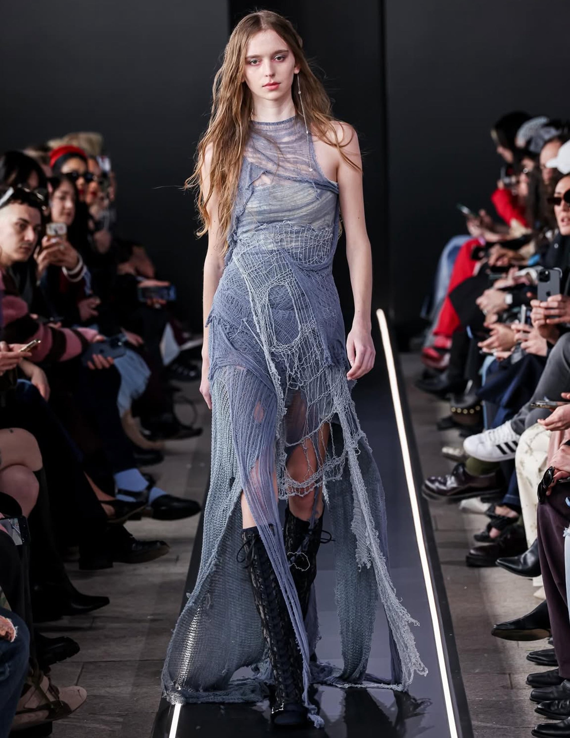 Model wearing a blue, intricately designed dress on a runway with an audience.