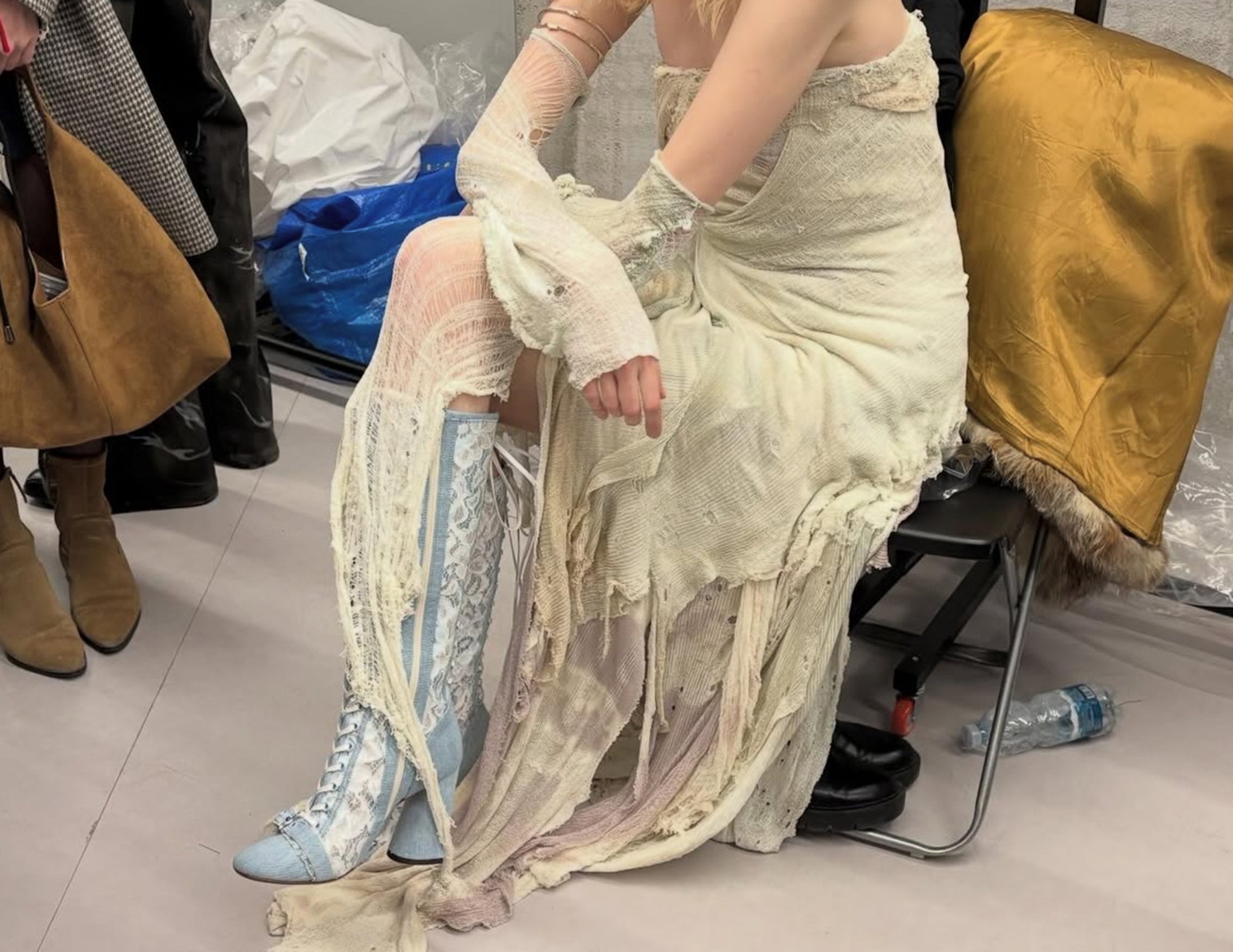 Person wearing a sheer dress and blue boots sitting on a chair with others around.