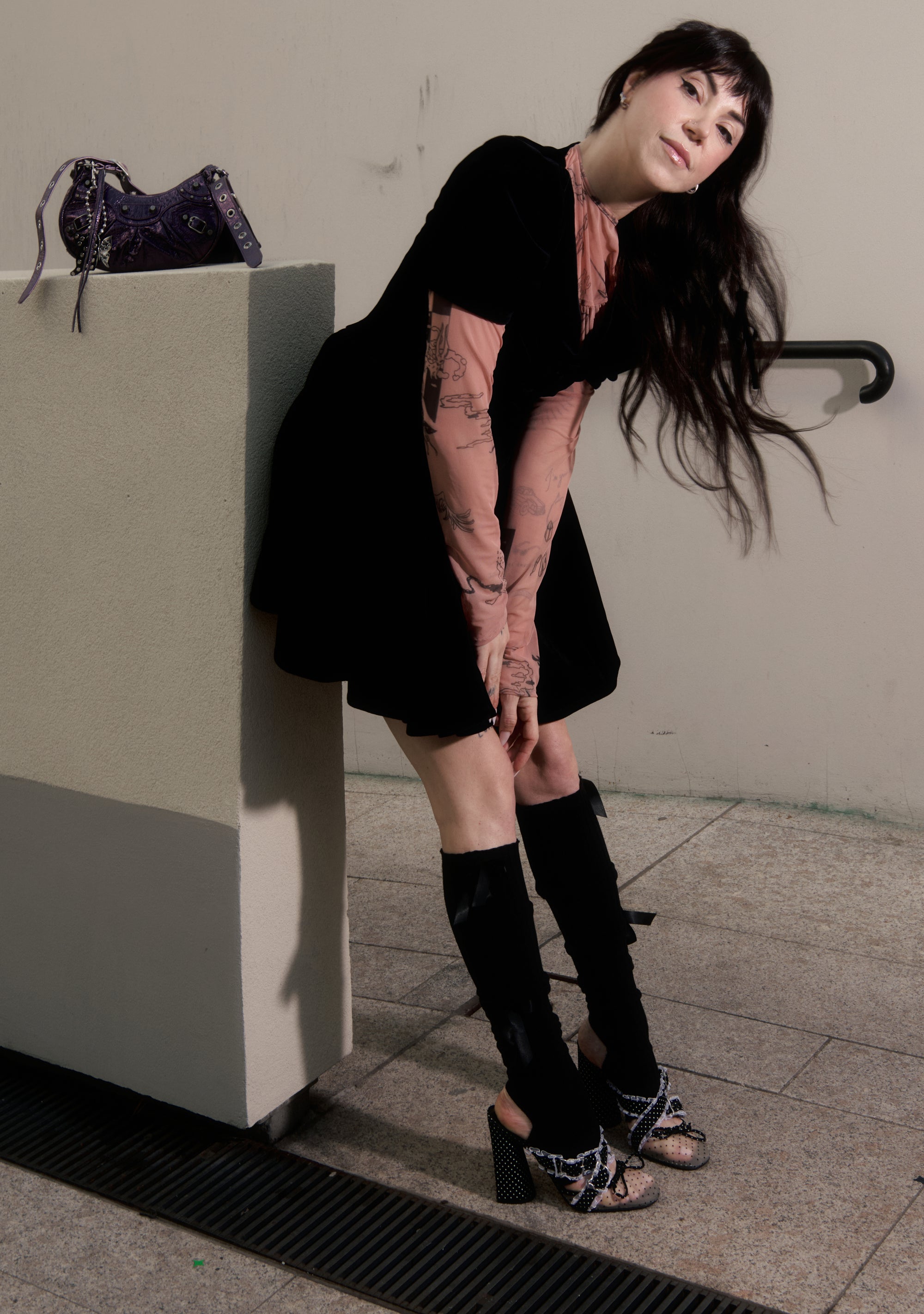 Woman in black dress and knee-high boots leaning against a wall with a handbag on a ledge.